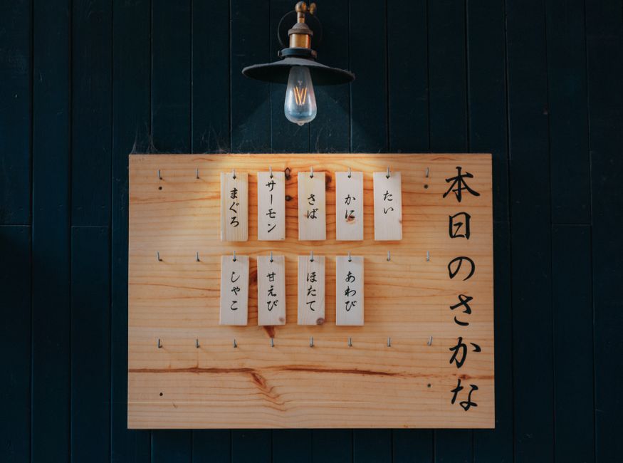 暗い壁の前に日本語のテキストが書かれたヴィンテージの木製メニューボードの上に灯った電球が吊るされています。