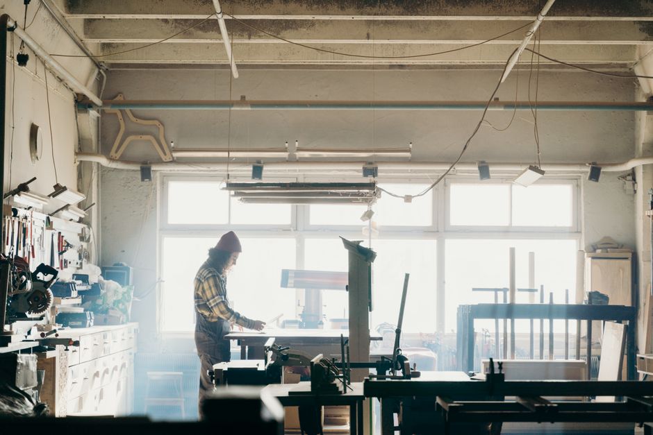 職人が日差しの差し込む工房で木工に集中しており、周囲には工具や機械が並んでいます。